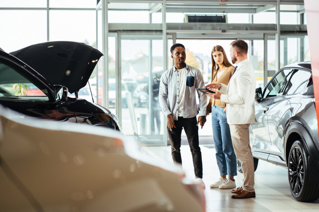 Competent car dealer using digital tablet for work with clients at showroom. Happy multiracial couple talking with manager while buying car at dealership center.