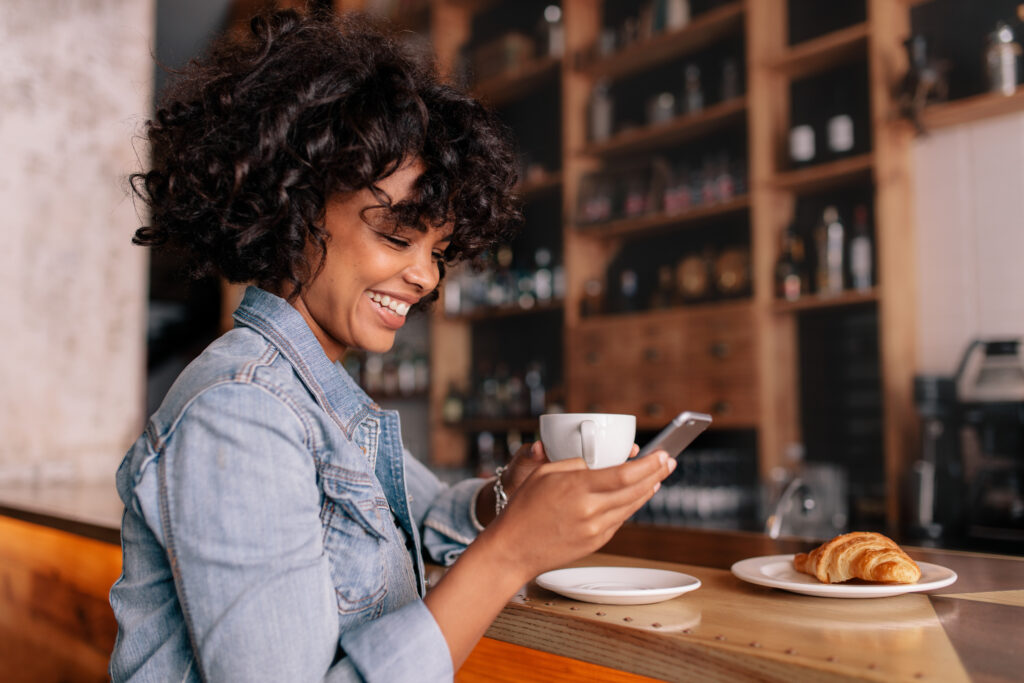 Smiling woman using smart phone in a modern cafe. Young african female sitting at a cafe counter having coffee and reading text message on mobile phone.