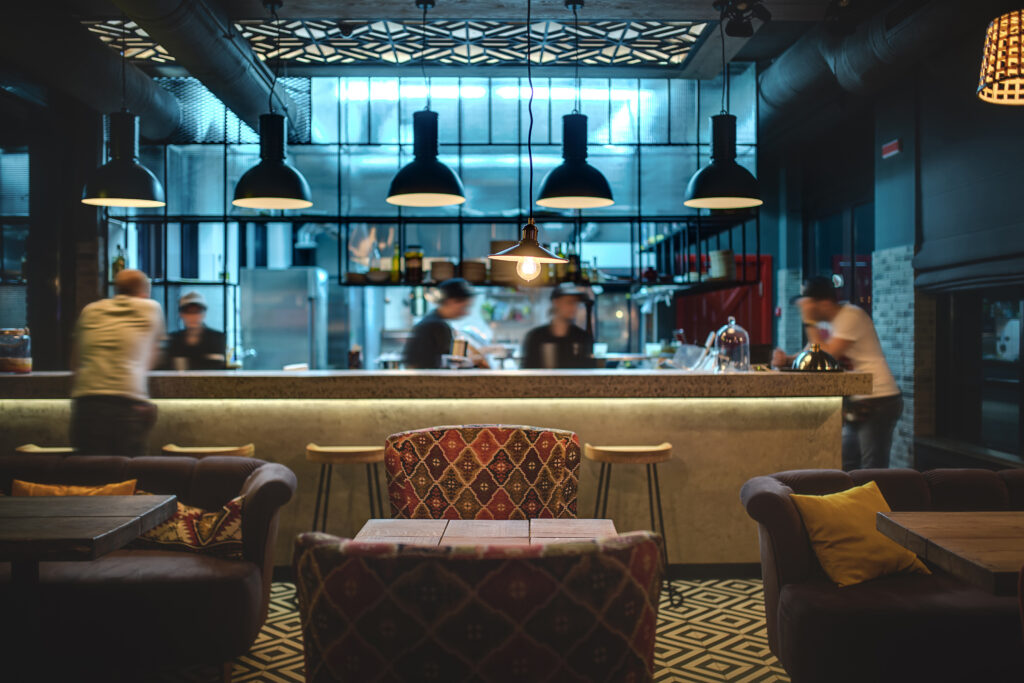 Half-lighted hall in a loft style in a mexican restaurant with open kitchen on the background. In front of the kitchen there are wooden tables with multi-colored chairs and sofas. On the sofas there are color pillows. In the kitchen there is a rack with wooden chairs near it, shelves with dishes and accessories, kitchen equipment. On the right there is a red wooden door, a brick wall with windows with curtains. At the top there are different glowing lamps and communications. On the floor there are black and white tiles with patterns. Horizontal.