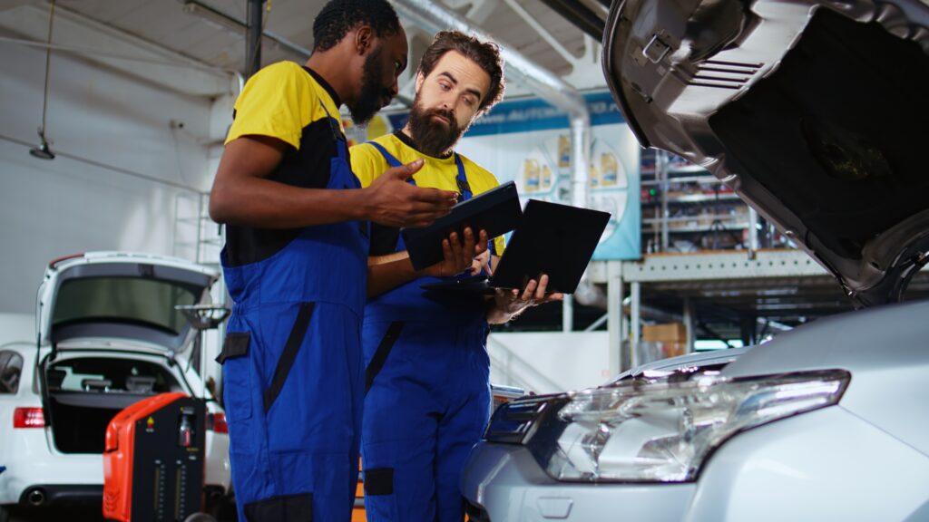 Repair shop employees work together on fixing car, using tablet and laptop. Teamworking colleagues collaborating on servicing broken vehicle, checking for defective components using electronic devices